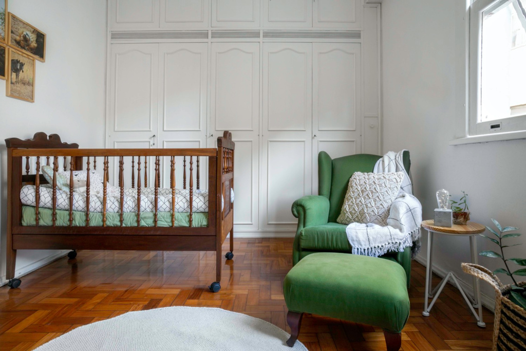 Chambre avec armoire blanche à l'arrière, lit bébé en bois foncé avec roulette, fauteuil vert et repose-pieds, à côté d'une petite table avec plateau rond et parquet.