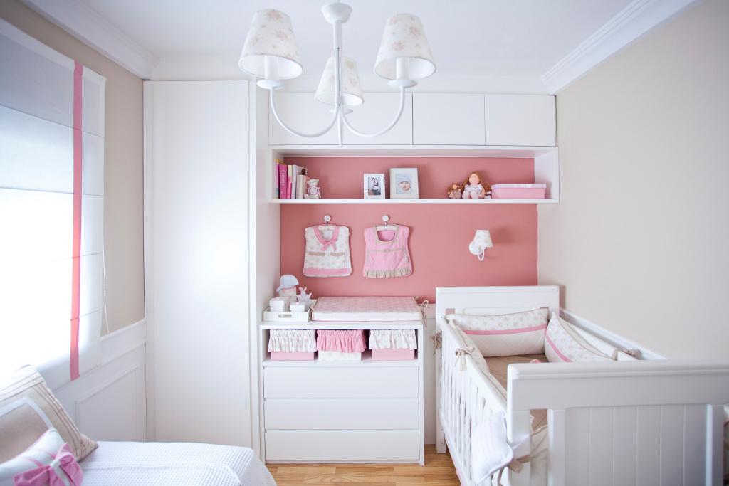 Petite chambre avec lit bébé et lit blanc.  Sur le mur, sur le côté gauche, une armoire jusqu'au plafond, à côté d'une commode avec un changement devant un mur rose, avec une étagère et une armoire à quatre portes à côté du plafond.