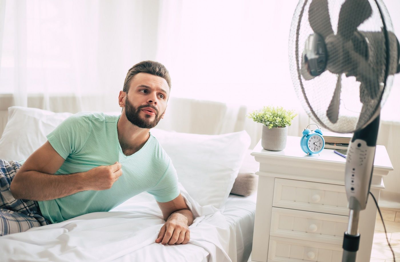 Un jeune homme en sueur essaie de se rafraîchir de la chaleur avec un ventilateur en position couchée dans son lit à la maison.