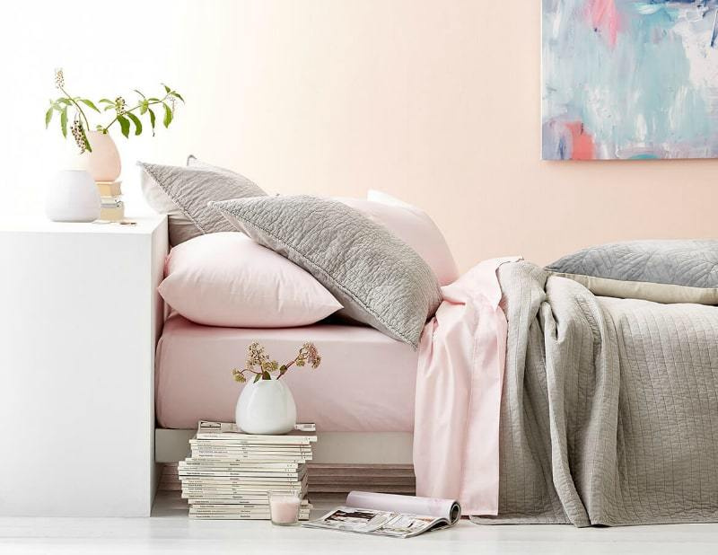 Chambre avec lit bas avec drap rose clair et oreillers gris.  Tête de lit blanche avec vase soutenu.  Pile de magazines avec petit vase sur le dessus