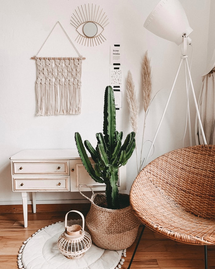Coin d'une pièce avec une chaise suspendue au plafond, un gros cactus dans un panier de paille, sur un tapis au crochet.  Appuyé contre le mur, un meuble blanc rustique est suspendu, un morceau de macramé blanc