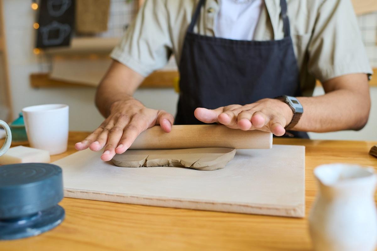 Mains d'un jeune homme noir en tablier aplatissant un morceau d'argile avec un rouleau à pâtisserie à bord tout en étant assis à table avec des fournitures pour le travail manuel.  Les différents types de pâte à modeler.