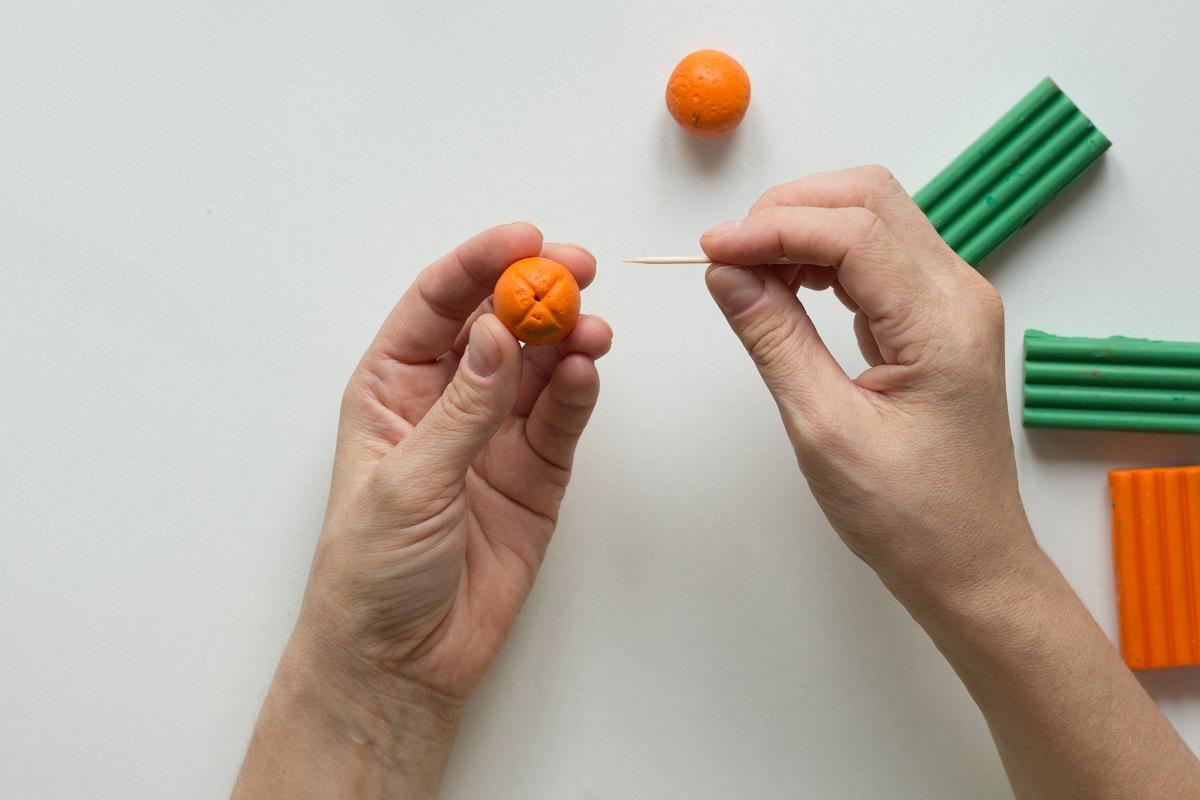 Mains de femme montrant un point sur une boule orange faite par un bâton dentaire pour créer un fruit orange à partir de l'argile polymère.  Pouvez-vous cuire des pâtes à modeler de tous types.